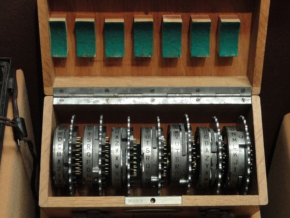 Device from an exhibit in the National Cryptologic Museum. A wooden box, opened on a hinge, displaying seven gears with the letters of the alphabet on them. This was likely used in conjunction with some kind of cipher system for code breaking.