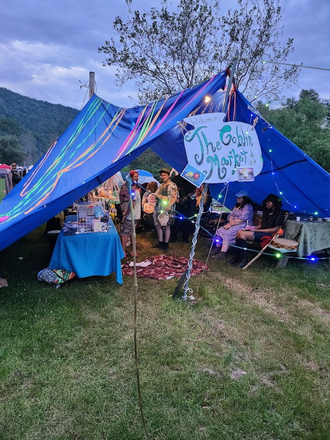 A blue tent decorated with colorful streamers, fairy lights, and a sign saying "The Goblin Market." People are sitting underneath.