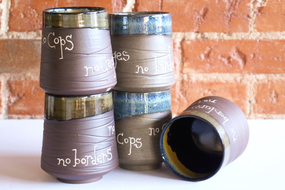 A stack of ceramic cups with inset foot rings and slightly conic walls. The outsides are mostly bare clay with overlapping rings of grooved texture, but the top inch or two around the lip is glazed at an angle, as well as the inside. Some are chocolate brown with a green and black glaze that turns gold inside the lip, and some a lighter speckled brown with speckled blue glaze. All of them have hand-carved white inlay lettering that says no cops, no cages, no borders.