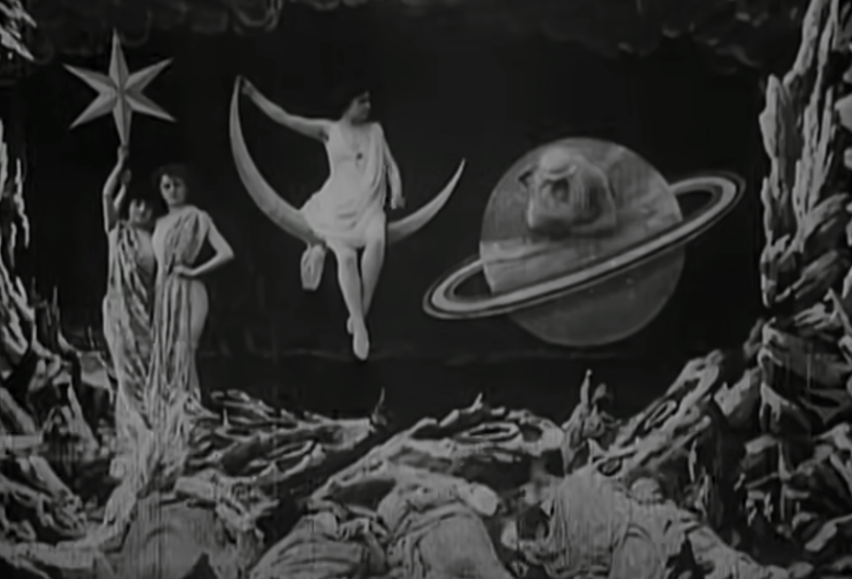 a black and white film still featuring actors posing with stylized celestial props like stars and a moon, on a theatrical lunar stage set