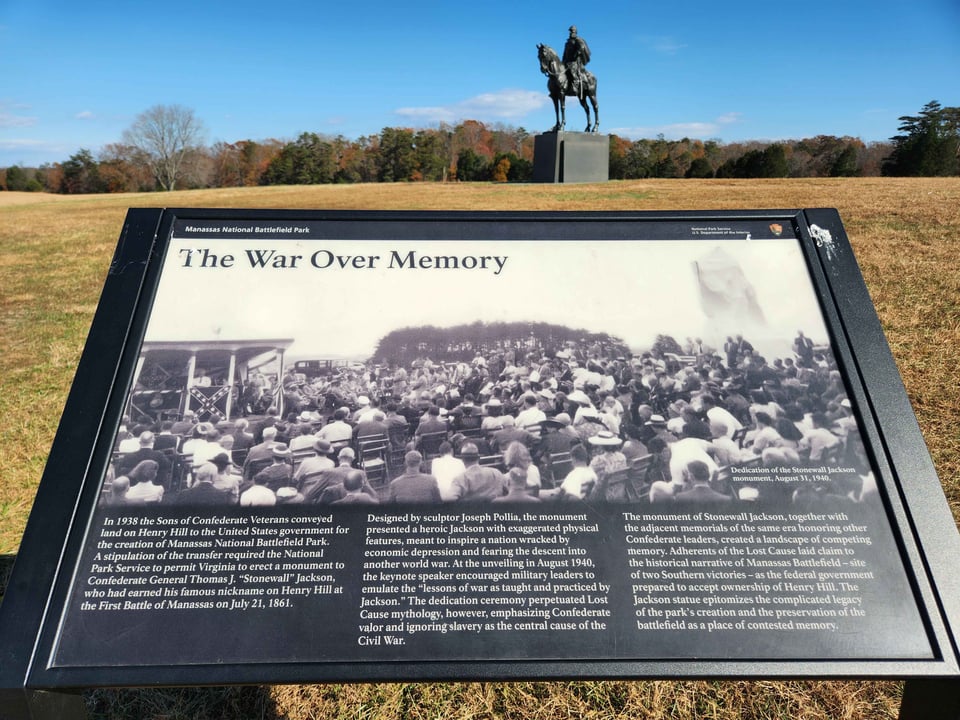 In the background, a statue of a man on horseback sits in a field under a blue sky. In the foreground, a plaque reads THE WAR ON MEMORY