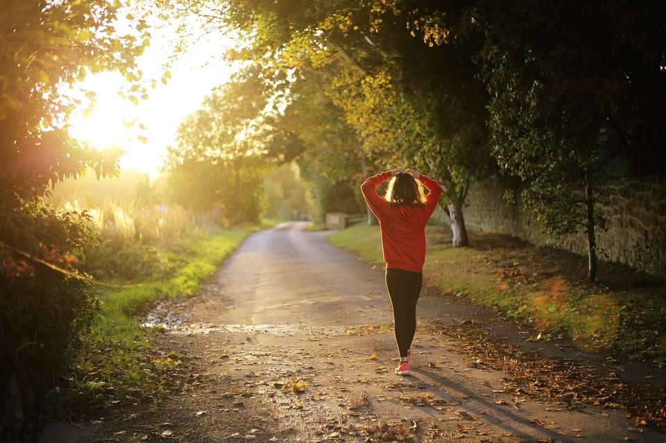 An image of a woman walking down a paved walking path with trees growing up on the right side. On the left is some scrub grass and the light of the sunset as the sun approaches the horizon.