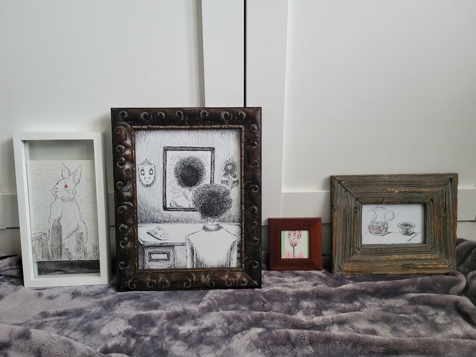 A row of four framed drawings. The first one is a drawing of an albino rabbit towering over the San Francisco downtown skyline. The second is of a person looking at themself in the mirror only to see a void instead of a face. The third is a very tiny drawing of red tulips within tulips and surrounded by flames. The fourth is a drawing of a pitcher of noodles next to a ramen bowl.