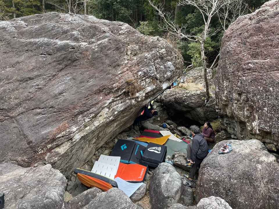 Amego boulder. Overhung, with a wealth of pads placed underneath. Two spectators sit and watch a climber working a hard sequence on the overhung side.