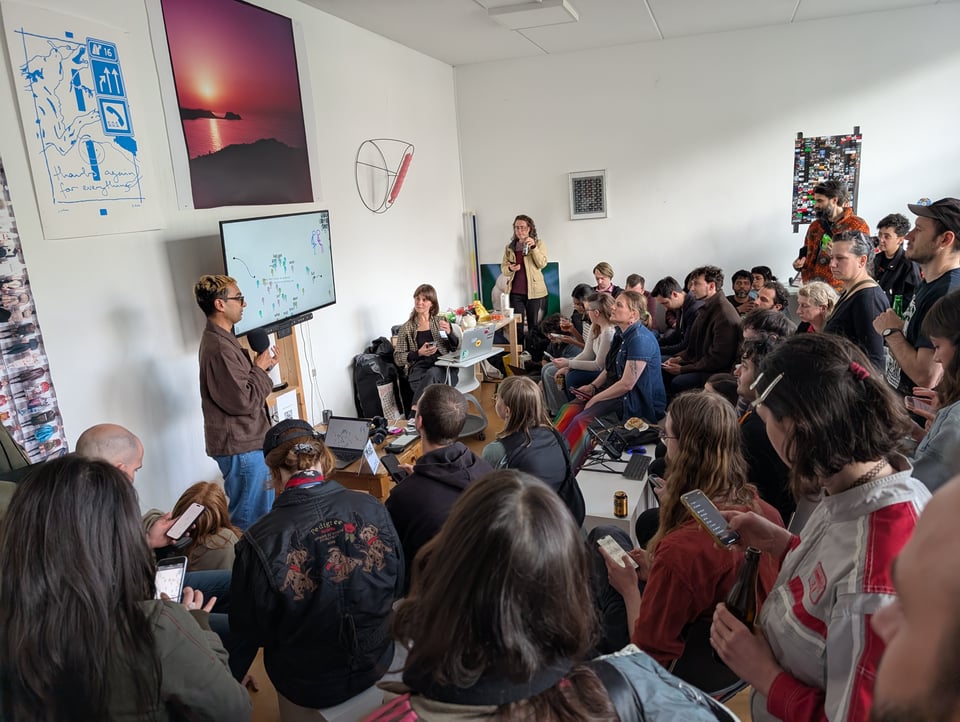 a speaker with a microphone address a room full of people, some seated, some standing. A TV stands in the center.