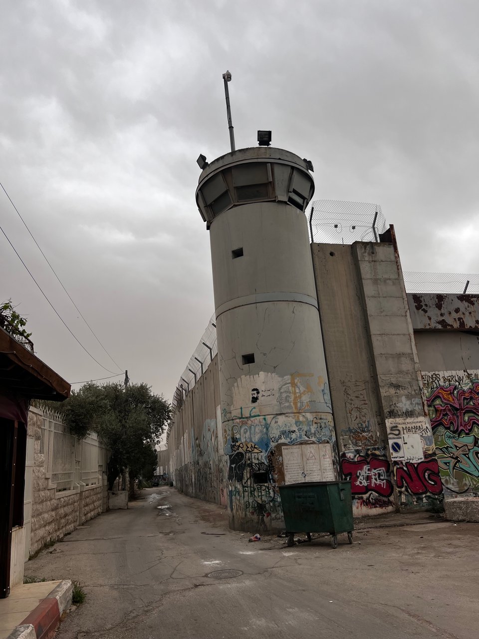 A large concrete wall topped with barbed wire on one side of a narrow street, and a tall guard tower with windows all around at the top