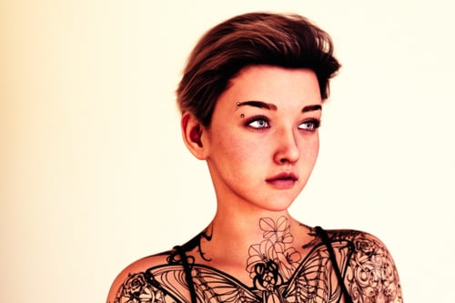 A portrait of a woman with short dark hair, against a bright plain background. She is looking off to the right, not at the viewer. She is heavily tattooed on her chest, shoulders, and arms. She has an eyebrow piercing on her right eyebrow.
