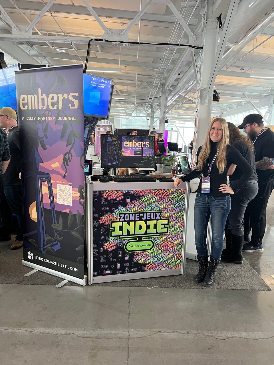 A white femme-presenting person with long blond hair stands next to a booth for a game called "embers: a cozy fantasy journal." They are wearing a black turtleneck and jeans.