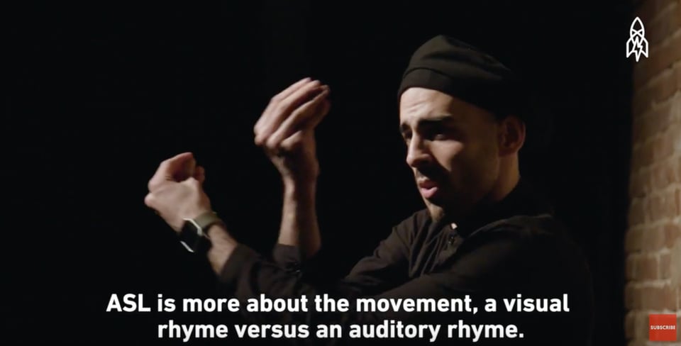 Screenshot of a YouTube video showing the upper torso of a man facing to the left. His hands are cupped and he is gesruring with them towards his head. The lighting is very dramatic and has a chiarascuro effect; he is facing a light that is out of frame, and his face and hand are brightly lit while his body -- a black cap and black sweater -- are muh less visible, and the backgrond is entirely black. White text appears at the bottom of the screen that reads: "ASL is more about the movement, a visual rhyme versus an auditory rhyme."