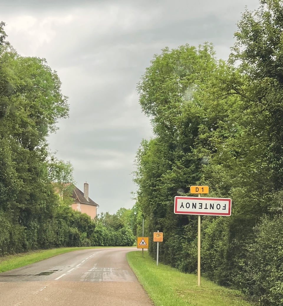 sign for village of Fontenoy upside down with slightly wet road and village in background, surrounded by wood