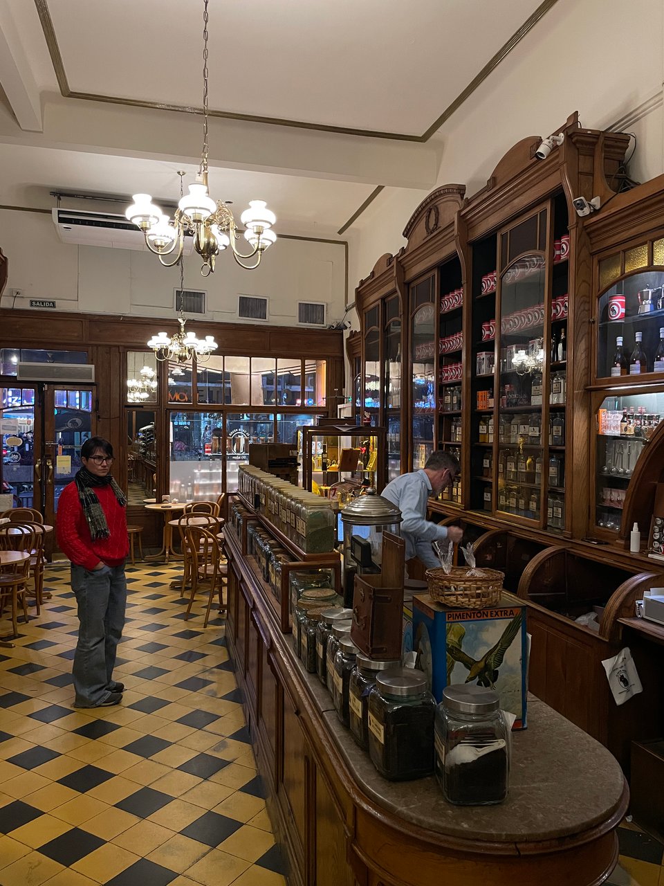 A woman in a red sweater looks at the counter full of teas in glass jars. A man working behind the counter is busy sweeping the floor.
