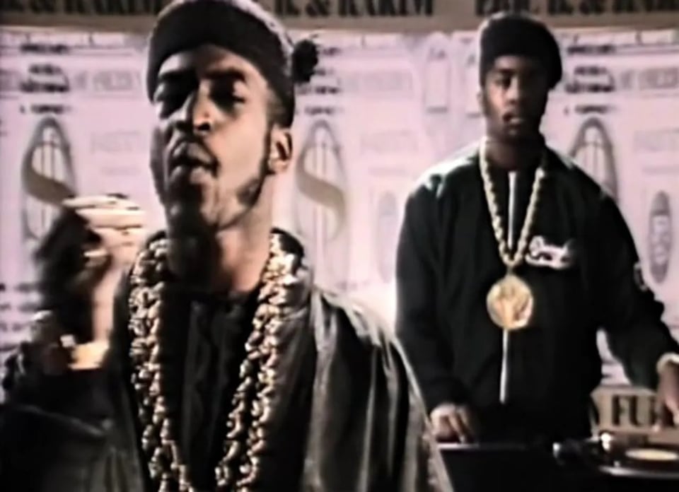 A still of Eric B and Rakim from the video for "Paid In Full." Eric and Rakim both have track suits, kangols, and gold chains. Rakim is making a "money" gesture" while Eric B spins turntables behind him.