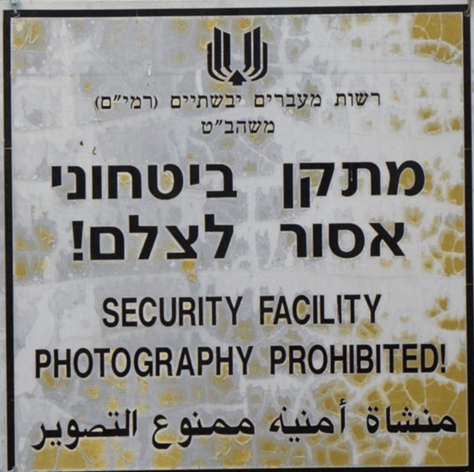 A photograph of a sign with large Hebrew writing, followed by the words in English:  SECURITY FACILITY / PHOTOGRAPHY PROHIBITED!  And then Arabic writing at the bottom