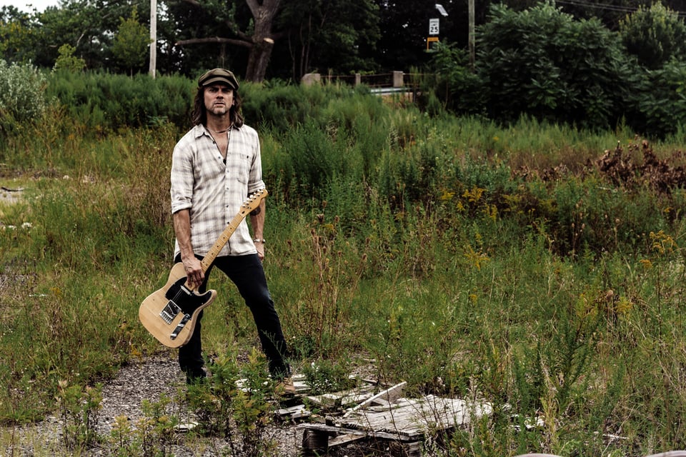 A man in a cap and a white-checked flannel shirt stands holding a blond Fender Telecaster guitar in a weedy lot.