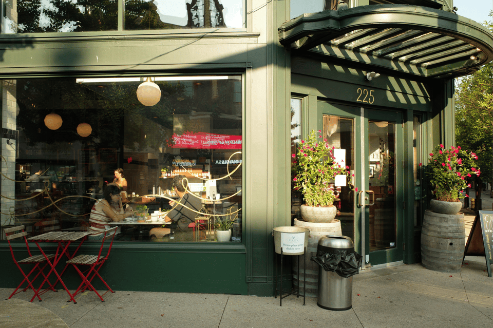 Building back when it used to be Ellie's. Dark green trim and glass storefront, small red café tables on the sidewalk, and people sitting inside by the window.