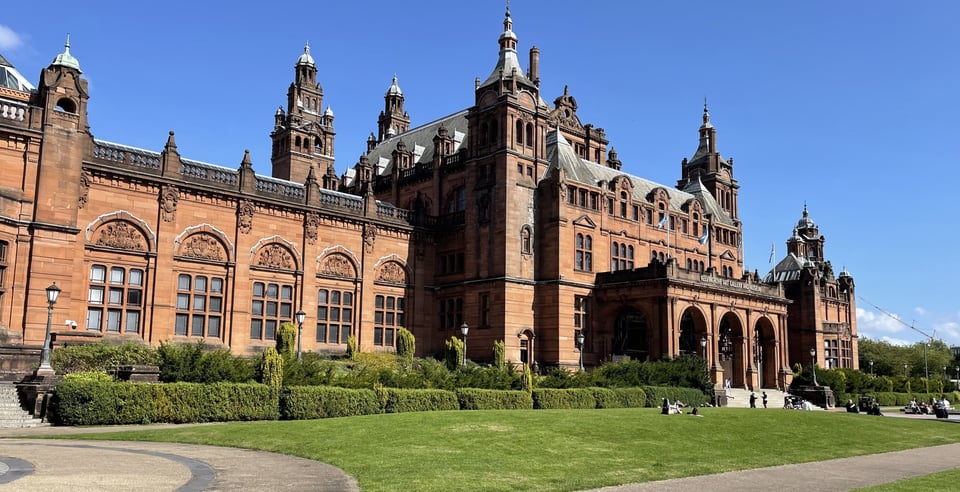 A picture of the exterior of the Kelvingrove Art Gallery and Museum - an ornate red sandstone building against a bright blue sky and green grass.