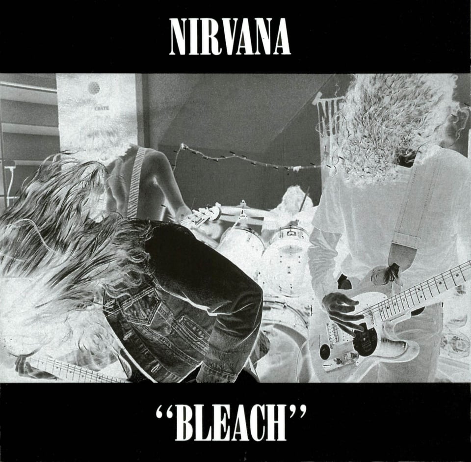 A black and white negative (creep) image of Nirvana playing (three guitarists and one drummer) all of them banging their heads and making their hair whip around.
