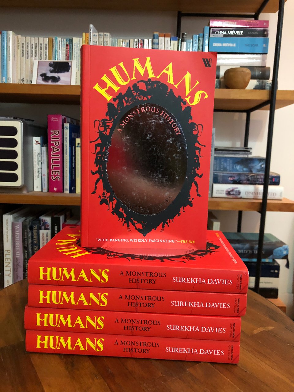 A tower of copies of HUMANS, with bright red spines, and a single copy balanced on top, its mirror-foil oval mirror on the cover facing outwards.