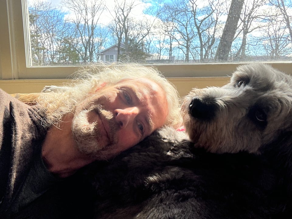 photo of white man with long hair resting his head on a gray aussiedoodle; she is looking away and rolling her eyes at the camera