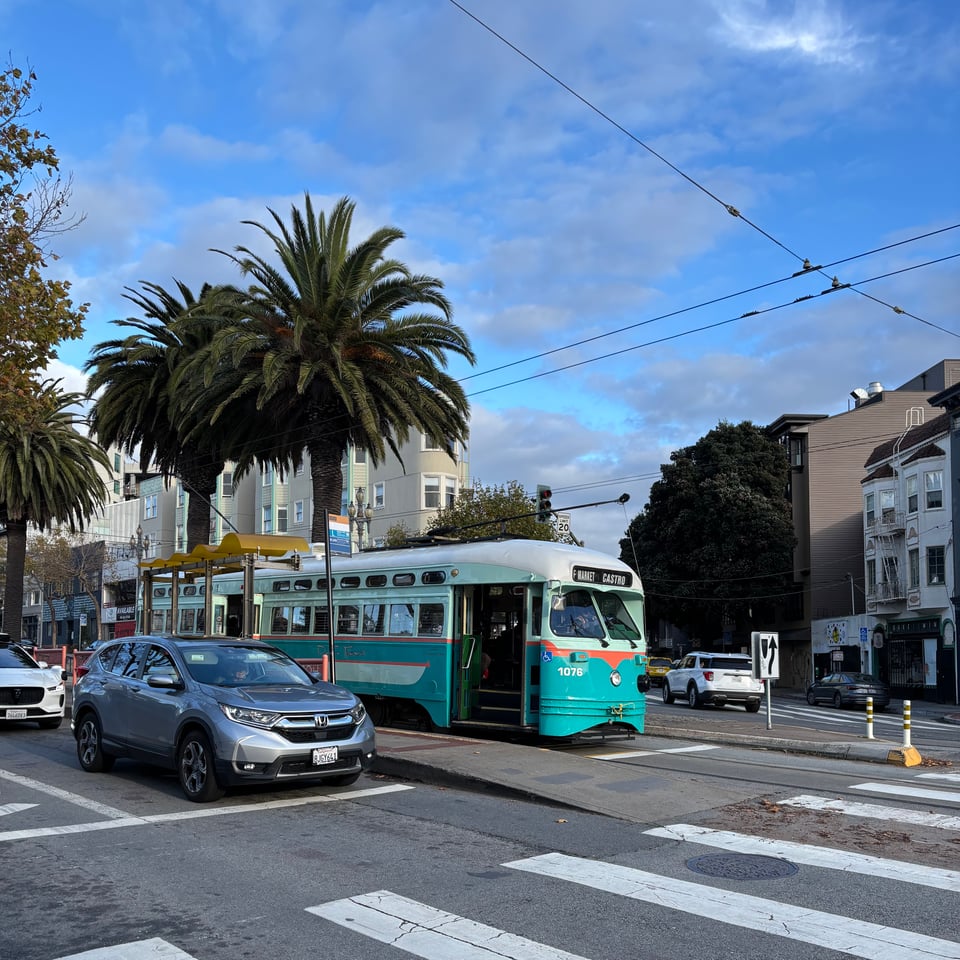 A teal and orange streetcar sits alongside a lane of cars at a San Francisco crosswalk. There are palm trees between the lanes, and grey apartment buildings in the background.