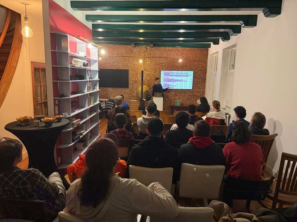 A man stands at a podium with a screen behind him, and speaks to the audience sitting in the room in front of him.