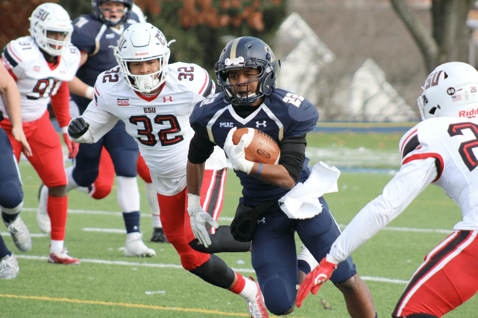 A photo of a football game. A man in a black uniform is holding the football and running, while two other players in red and white uniforms prepare to tackle him.