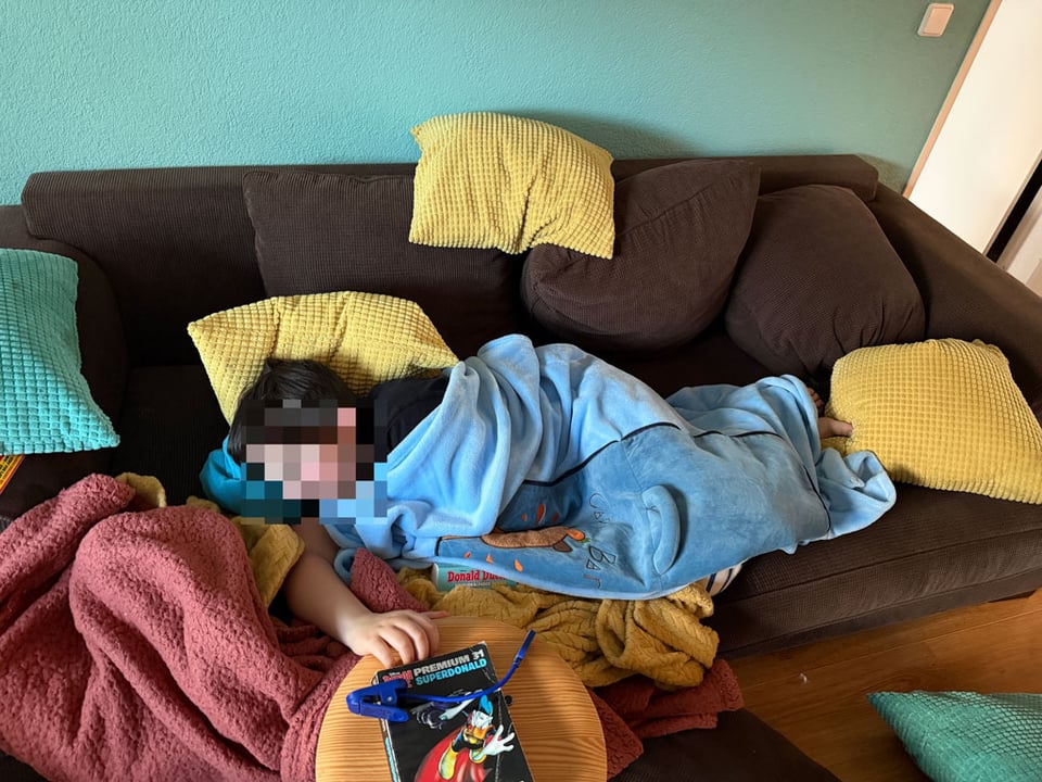 A young boy sleeps on a couch surrounded by pillows and blankets and books.