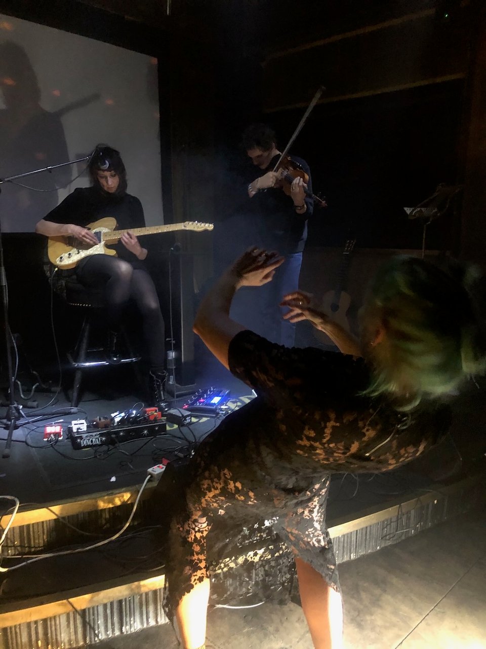 Alison Clancy on stage at Gold Diggers, playing guitar. She's accompanied by a violinist and a dancer who is on the floor in front of the stage and bending backwards