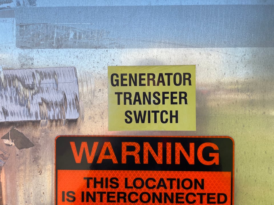 Two signs posted on an electrical box in my hometown. The first one is yellow and reads "GENERATOR / TRANSFER / SWITCH". The second is orange and reads "WARNING / THIS LOCATION IS INTERCONNECTED". Photo by me, 2023.