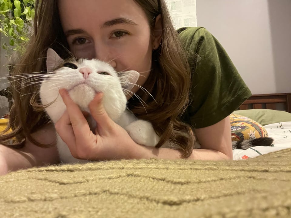 Copeland and their cat Anya lying on a bed with a green throw blanket beneath them. Anya is lying between Copeland's arms and Copeland is scratching Anya's chin
