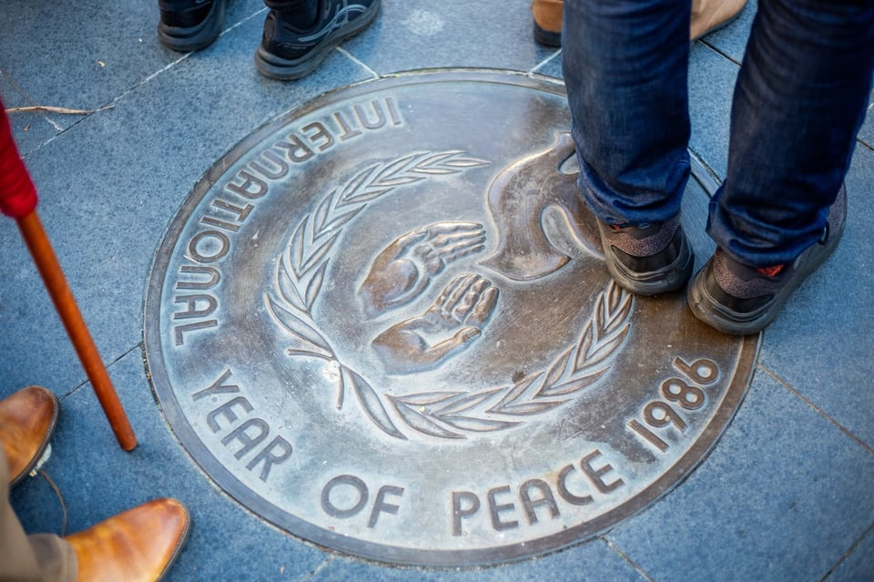 A bronze plaque with a picture of two hands releasing the dove of peace surrounded by two laurel branches. The text reads 'International Year of Peace 1986'. A number of feet are visible standing around the plaque.