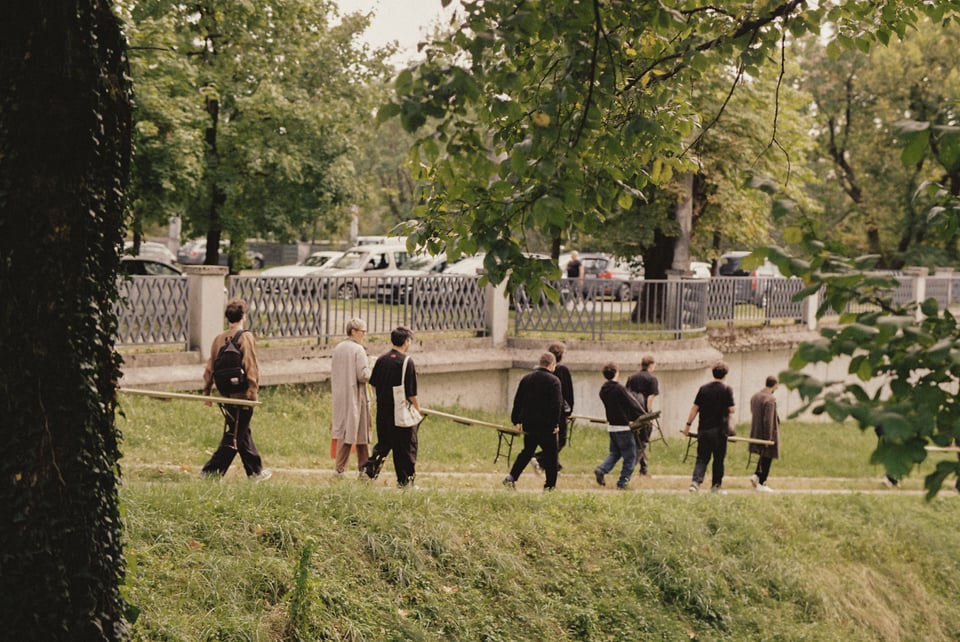 [Pairs of people hold either end of several long wooden benches and walk down a path in single file.]