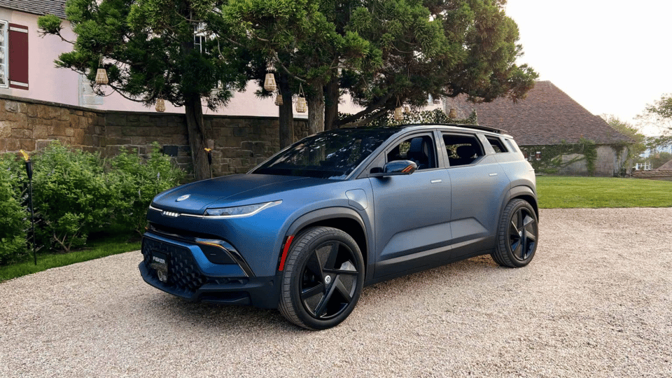 A picture of blue electric car styled very much like a small SUV, parked on a gravel driveway in front of a old fashioned house made of stone brick, with two trees behind