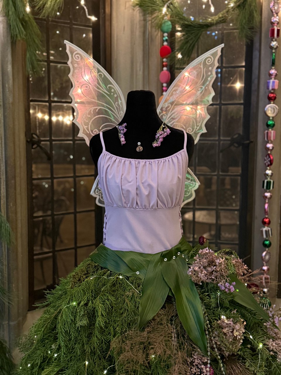 photo of pale pink bodice on a headless/armless black mannequin, with gossamer filigreed wings, and a skirt of evergreen branches and flowers, in a Christmas-themed manor room. Lights strung up outside through the window appear to be illuminating the wings.