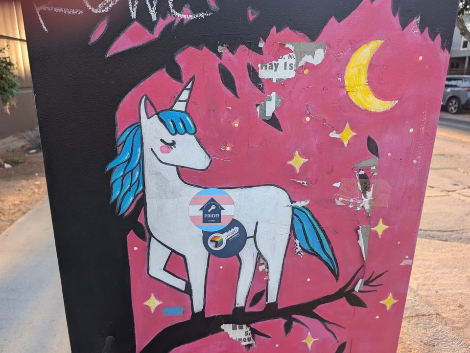 this street art has a pink background with a crescent moon and stars. A unicorn with blue hair and tail sits on a dark tree branch. It has a pink blush on its face. THere is a trans pride sticker on the unicorn.