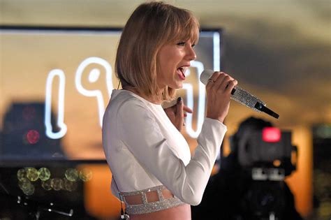 Taylor Swift singing into a microphone on stage. There’s a TV camera behind her (I think?) and a sign that reads 1989.