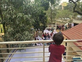 A small boy with dark hair looks out over a balcony on to a gathering of people. Lush green trees are visible, and white chairs with people gathered below.