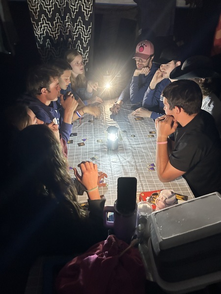 Nine teenagers and young adults sit around a camp table, playing a game in the dark, using a lamp for light.