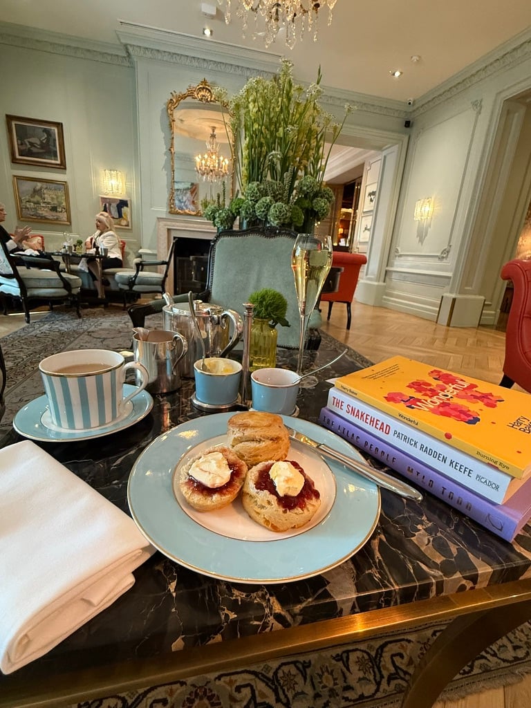 A table set for afternoon tea in an elegant pale-blue room, with tea, scones topped with jam and cream, a glass of sparkling wine, and a stack of books beside them.