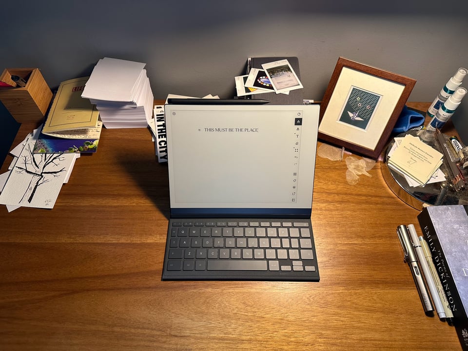 Photo of a desk with a reMarkable tablet + type folio at the center. Elsewhere there is a notepad, a stack of blank notecards, pens, postcards, Instax photos, a framed print of a swan, and a copy of Emily Dickinson's poems
