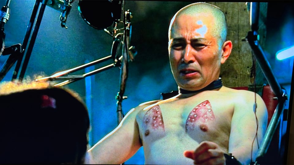 The abducted CEO in Save the Green Planet, cuffed and bound to a chair, shaved bald, shirtless, with burn marks from a steam iron on each of his pecs.