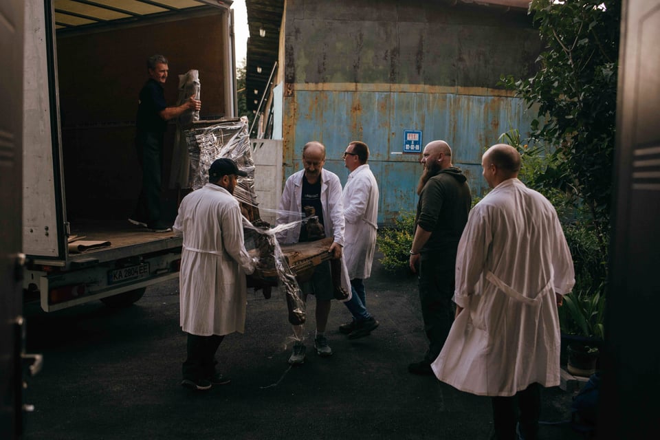 Ukrainians in white coats and black hoodies move a large chair from a white truck onto the ground.