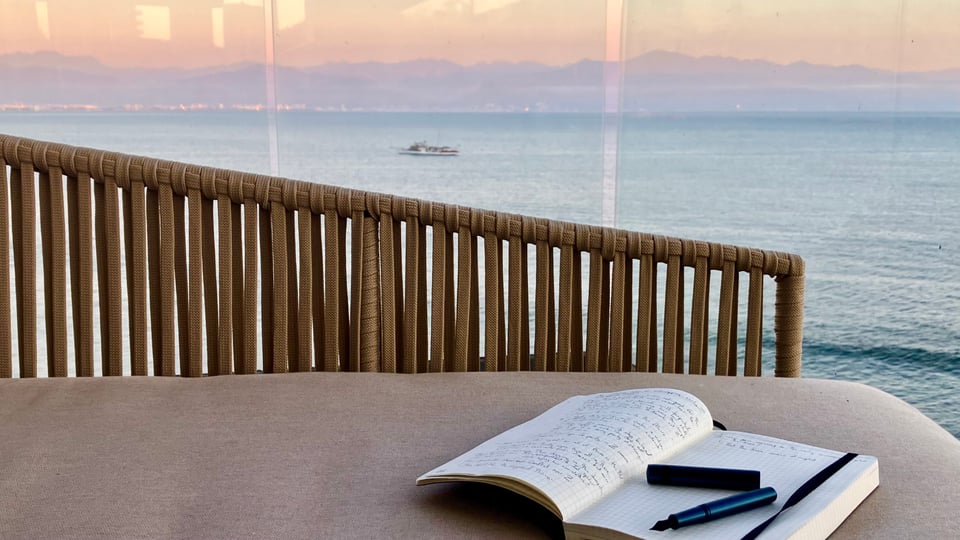 A notebook with messy handwriting sits open on a beige peice of outdoor furniture. A fountain pen sits uncapped on top of it. There is a sunset view of the ocean past the glass behind the furniture.