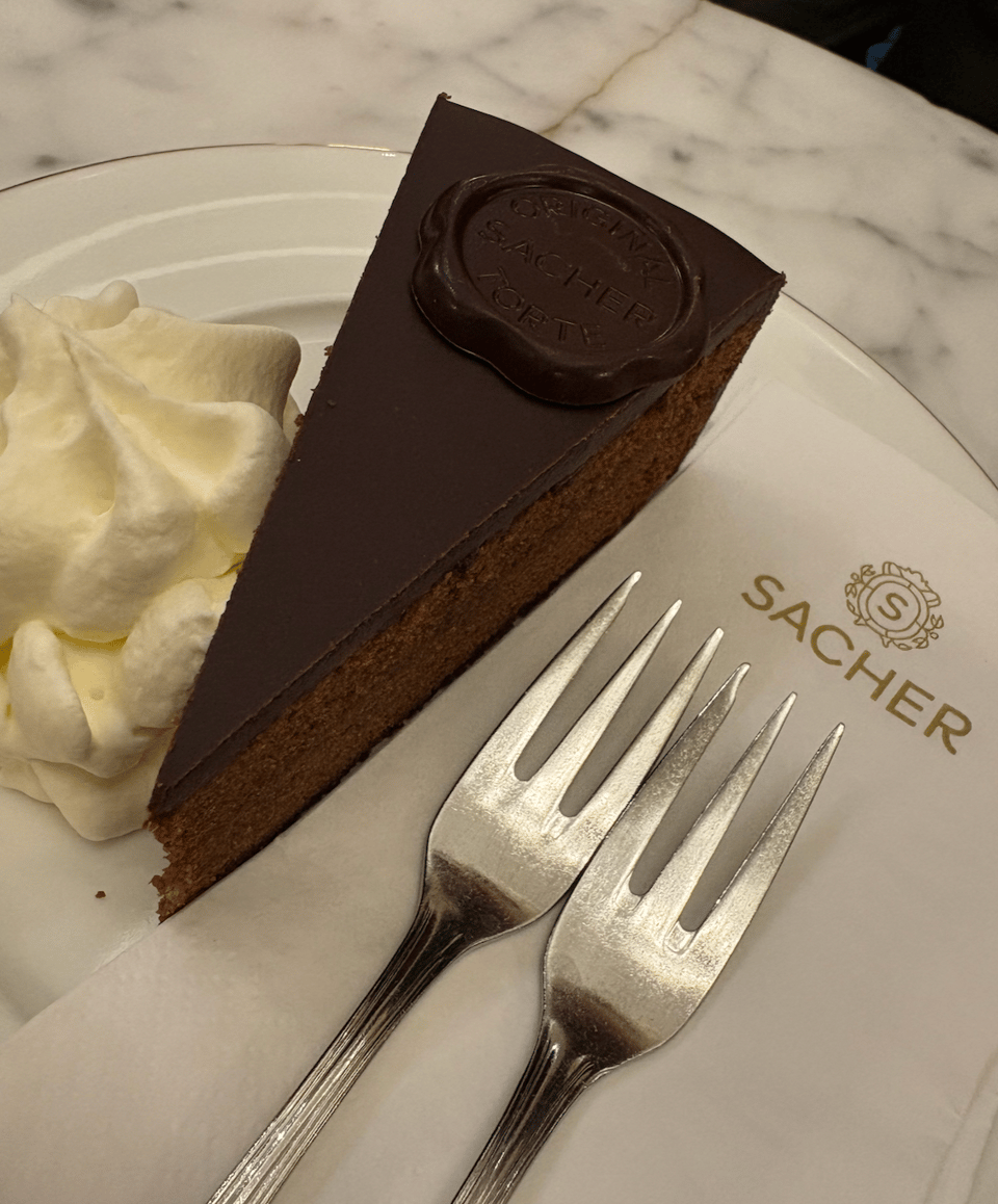 Photograph of a slice of a rich chocolate cake with a smooth dark chocolate glaze and a chocolate seal on top, served with a dollop of whipped cream on a white plate. Two silver forks rest on a white napkin printed with the word SACHER in gold, and a gold logo with an S in the center