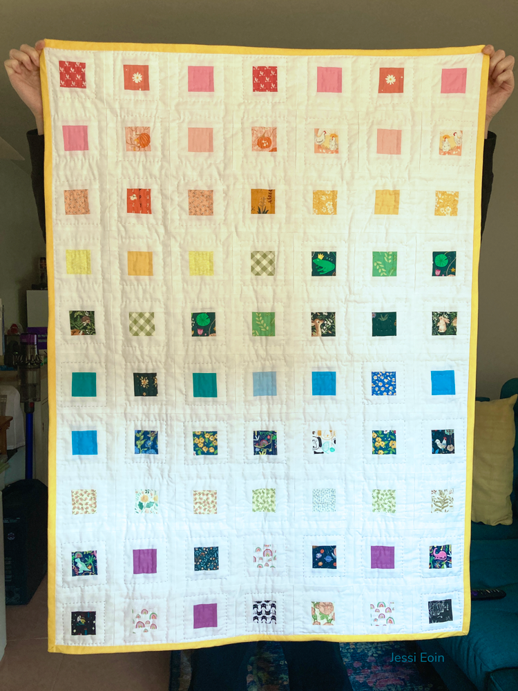 A tall person holds up a baby-sized quilt in front of them to show it off. The design shows colorful squares in a rainbow patten; the squares are made up of color scraps, tucked inside a larger white square, all of it tied together by a cheery yellow binding.