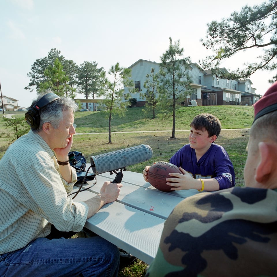Photo of John Biewen holding a microphone and sitting at a picnic table across from a boy holding a football, with a soldier in uniform looking on.