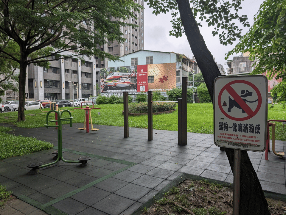 A small exercise area in a rainy park. There are uneven pullup bars and other machines whose function is unclear. In the foreground is a sign about not letting your dog poop there. In the background are a large apartment building and advertising billboard.