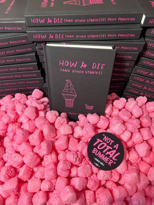 A stack of black books with the title How to Die (and other stories) by Mike Monteiro and cover pink line art of an IKEA soft serve cone full and eaten sits in a pile of pink heart-shaped packing peanuts. A round black sticker reading "Not a total bummer" - Erika Hall is in the foreground