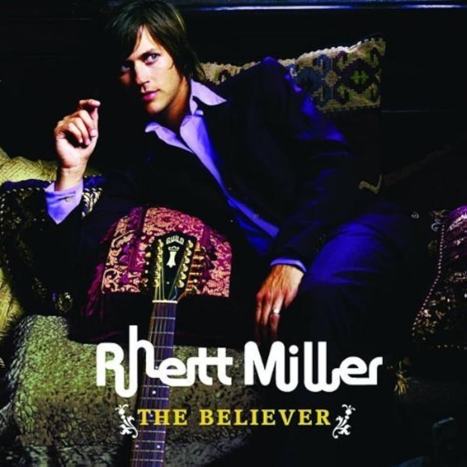 Album cover for The Believer by Rhett Miller. Miller is reclining on a patterned couch, dressed in a dark velvet suit with a light purple shirt. He rests his arm across a decorative pillow. The neck of a Guild guitar leans against the pillow beside him. The background is dimly lit, giving the image a moody, lounge-like atmosphere. The text “Rhett Miller” appears in bold white font across the bottom, with “The Believer” in smaller yellow type beneath it.