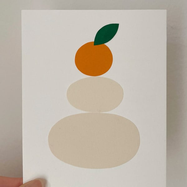 Postcard-sized artwork with cut paper shapes. One orange fruit stacked on two cream colored mochi oval circles